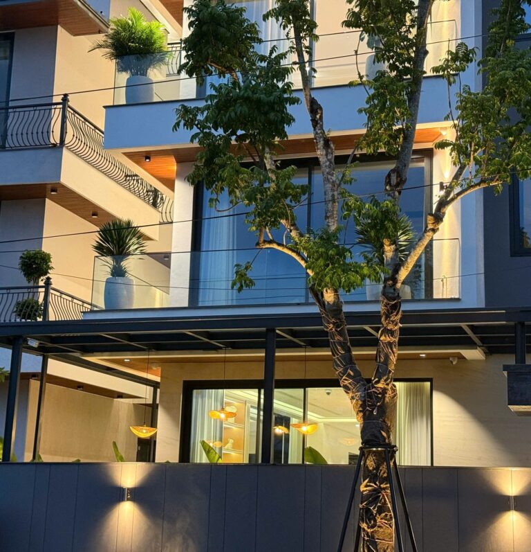 Mặt tiền biệt thự lúc chập tối: đèn hắt tường hai chiều, đèn rọi gốc cây và đèn thả mái hiên ánh sáng vàng ấm