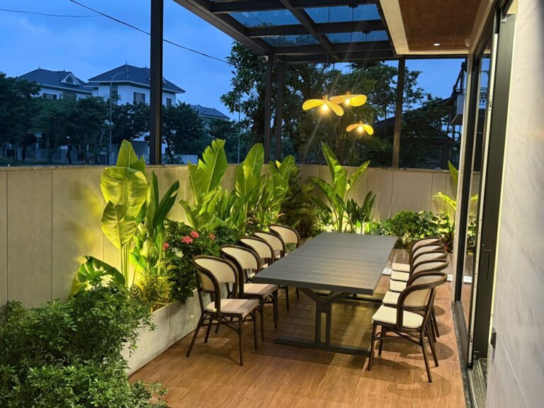 Sân thượng villa có giàn che kính, bàn ăn dài, đèn thả nón lá và đèn rọi cây xanh lúc chập tối