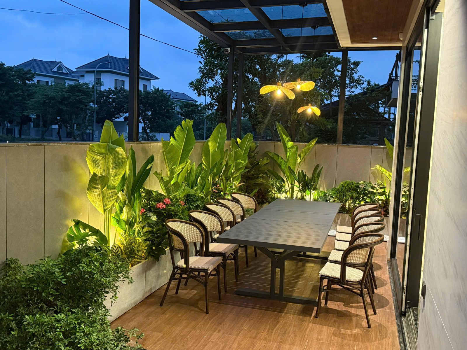 Sân thượng villa có giàn che kính, bàn ăn dài, đèn thả nón lá và đèn rọi cây xanh lúc chập tối