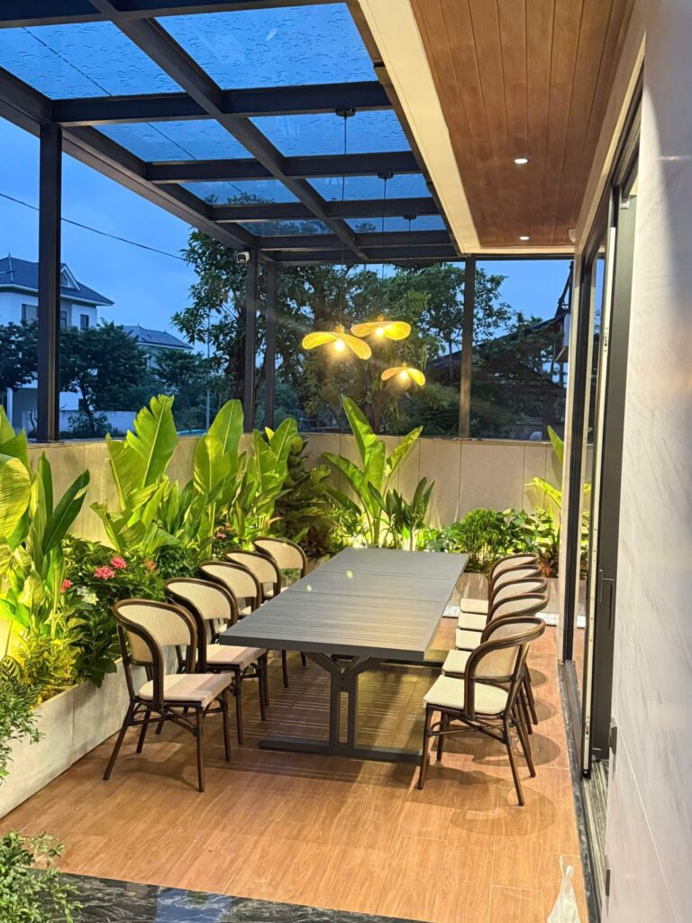 Khu vườn cây nhiệt đới trên sân thượng được rọi sáng từ dưới lên, đèn thả ánh sáng ấm phía trên bàn ăn
